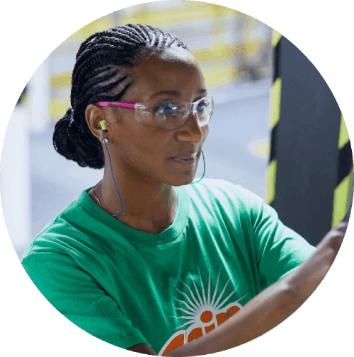 Woman with braided hair wearing glasses, earphones, and a green T‑shirt, standing in front of industrial equipment